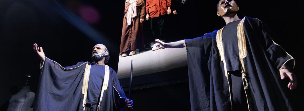 two actors in Classical Greek masks stand on a stage in front of a video screen showing women refugees from Syria